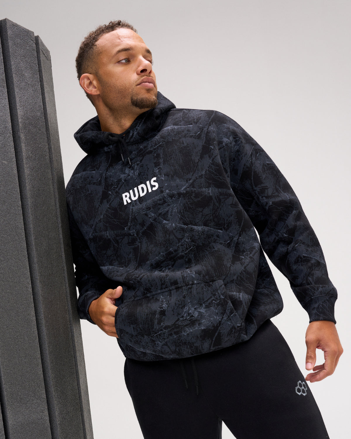 A man wearing a black and dark gray marbled hoodie with 'RUDIS' printed in white on the chest, leaning against a dark gray wall in a studio setting.