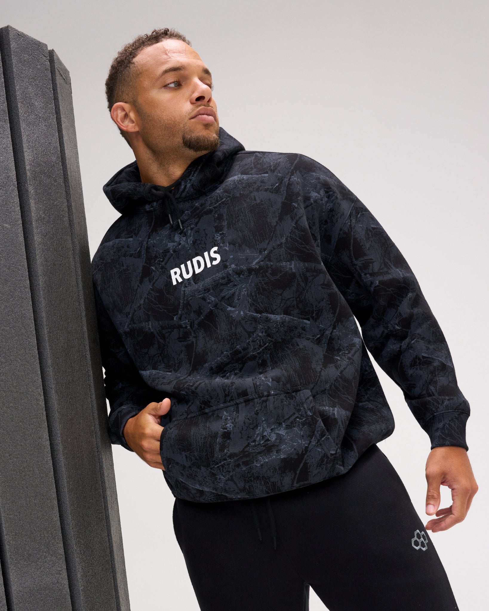 A man wearing a black and dark gray marbled hoodie with 'RUDIS' printed in white on the chest, leaning against a dark gray wall in a studio setting.