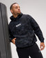 A man wearing a black and dark gray marbled hoodie with 'RUDIS' printed in white on the chest, leaning against a dark gray wall in a studio setting.