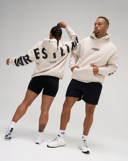 Two models wearing cream-colored hoodies and black athletic shorts, posed against a light gray studio background. The male model faces forward showing the 'FUDIS' brand logo on the chest, while the female model faces backward showing the back of the hoodie with 'WRESTLING' printed in black.