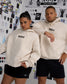 A man and a woman modeling matching off-white hoodies with the brand name 'RUDIS' printed in black across the chest, paired with black athletic shorts, posed in front of a white wall covered in fashion sketches and prints.