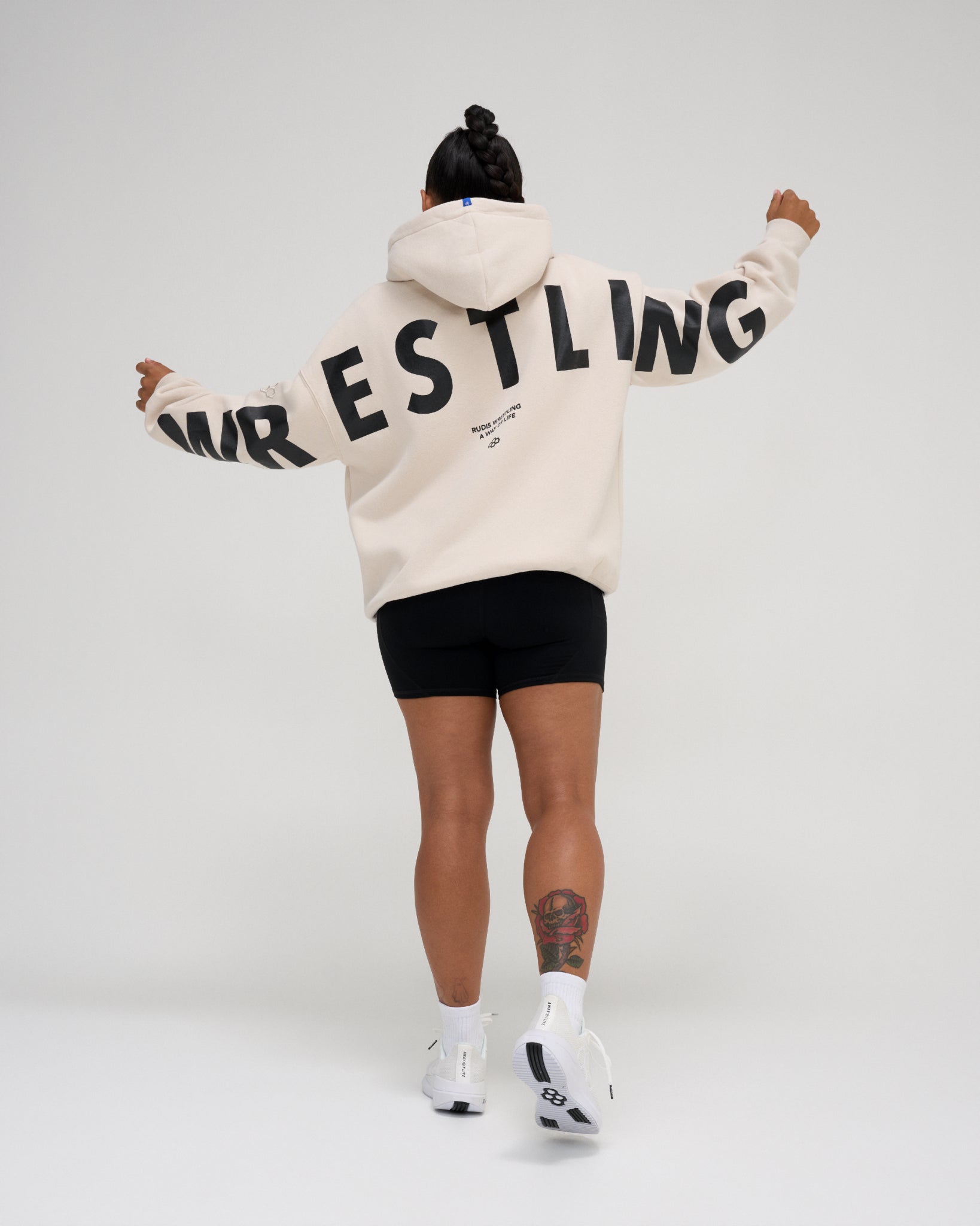 A person shown from the back wearing a cream-colored hoodie with 'WRESTLING' printed in bold black letters across the back, paired with black athletic shorts, white socks, and white sneakers, against a light gray studio background.