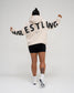 A person shown from the back wearing a cream-colored hoodie with 'WRESTLING' printed in bold black letters across the back, paired with black athletic shorts, white socks, and white sneakers, against a light gray studio background.