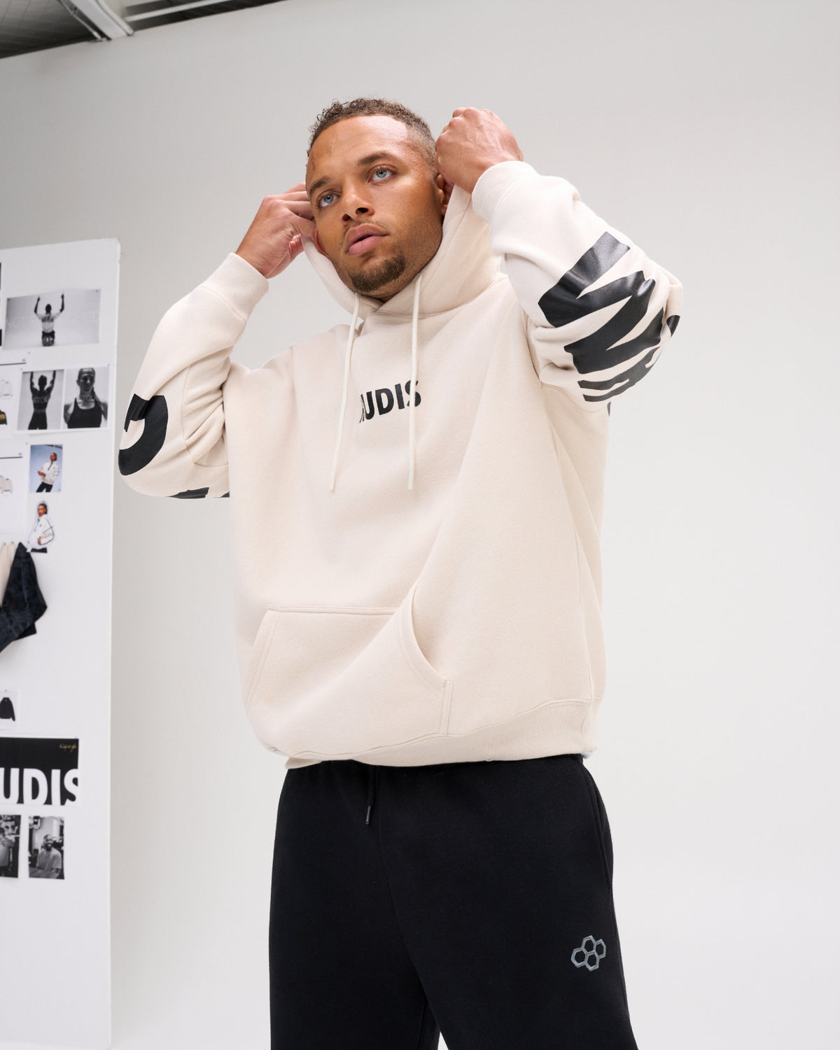 A man wearing a cream-colored athletic hoodie with black graphic details on the sleeves and chest, paired with black sweatpants, posing in front of a white studio wall.