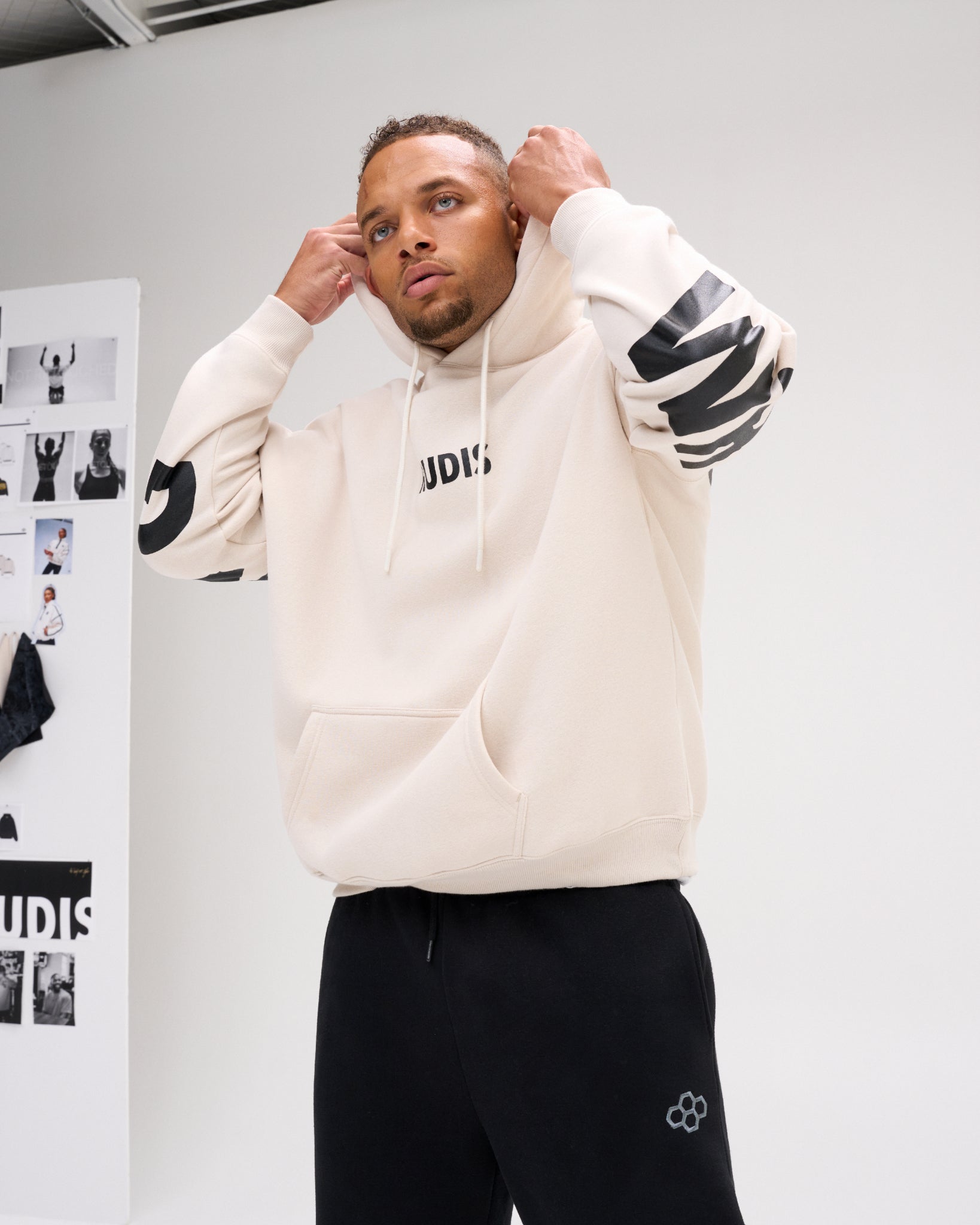 A man wearing a cream-colored athletic hoodie with black graphic details on the sleeves and chest, paired with black sweatpants, posing in front of a white studio wall.