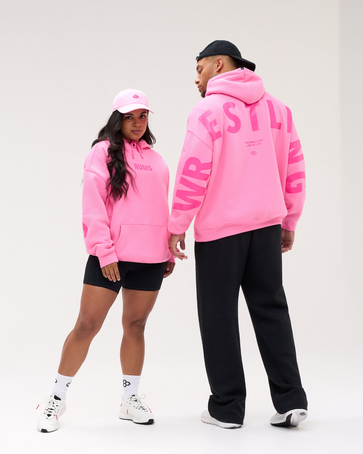 Two models wearing matching bright pink hoodies from the brand WRSTL, one shown from the front wearing a pink cap and the other from the back showing large pink lettering on the back, set against a plain white studio background.