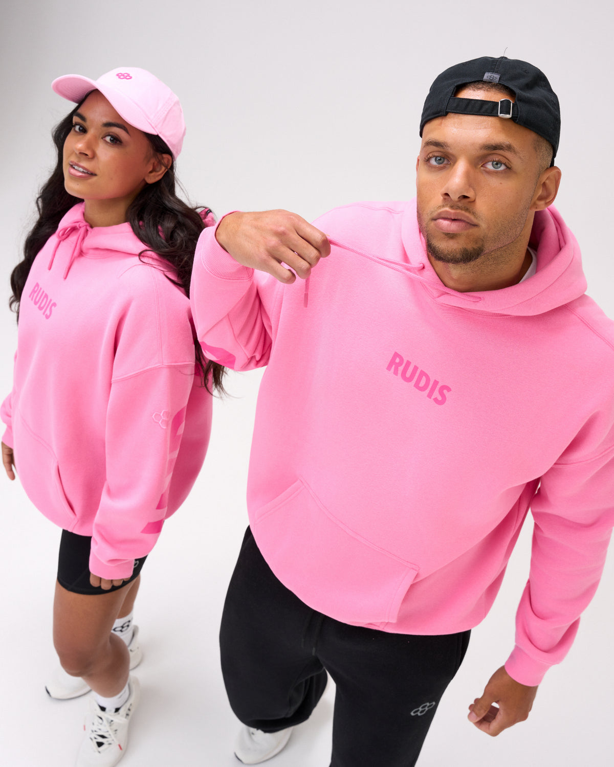A man and a woman modeling matching bright pink hoodies with 'RUDIS' printed on the chest, against a clean white studio background.