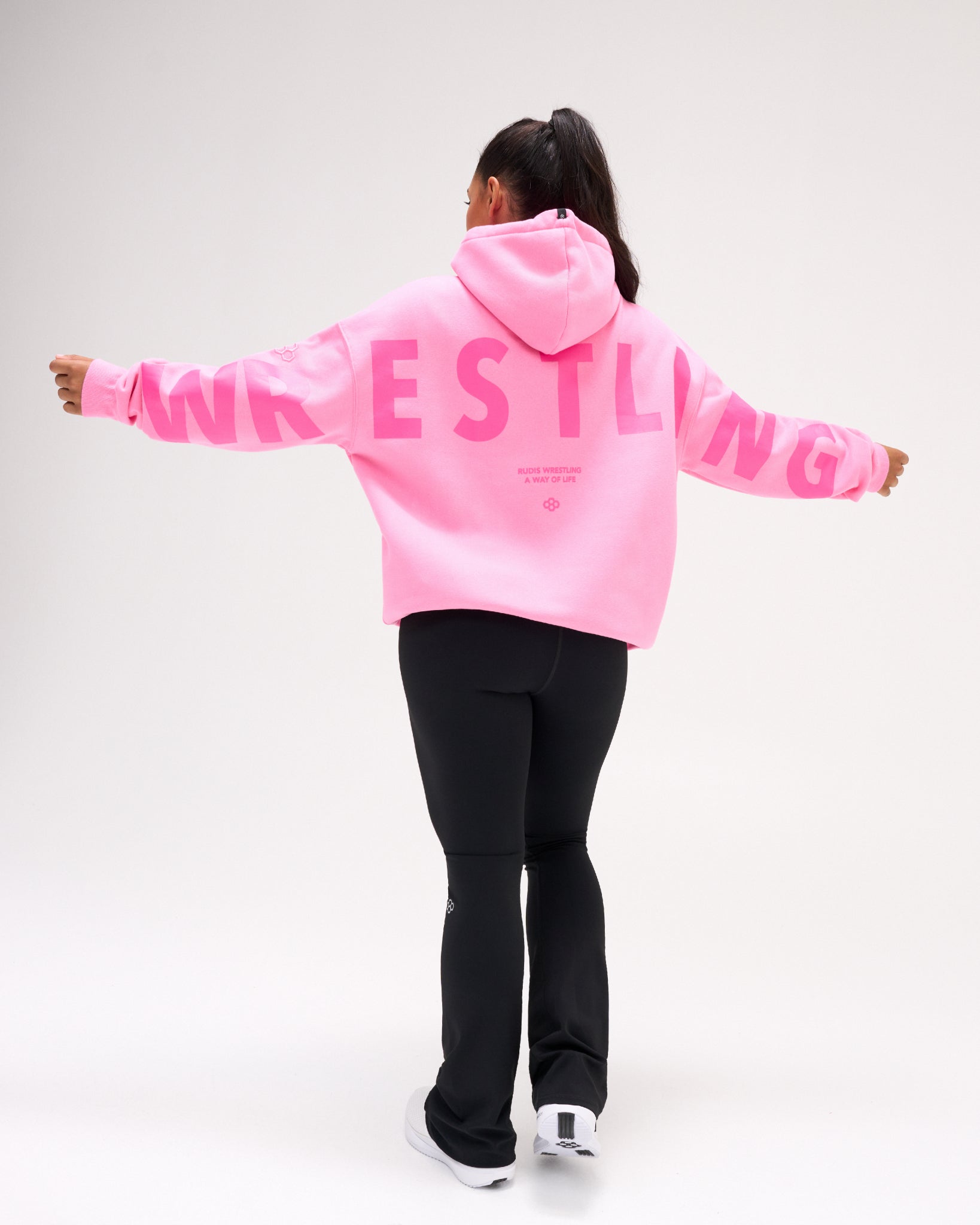 A model viewed from behind wearing a bright pink hoodie with 'WRESTLING' printed in large pink letters across the back, paired with black pants and white sneakers, set against a plain light gray studio background.