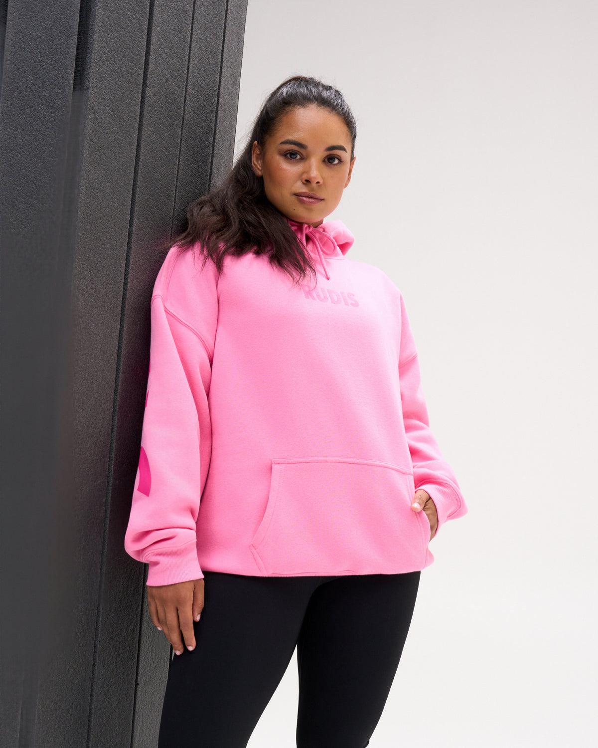 A woman wearing a bright pink hoodie with a tonal pink 'adidas' logo on the chest, paired with black leggings, leaning against a dark gray textured wall.