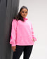 A woman wearing a bright pink hoodie with a tonal pink 'adidas' logo on the chest, paired with black leggings, leaning against a dark gray textured wall.