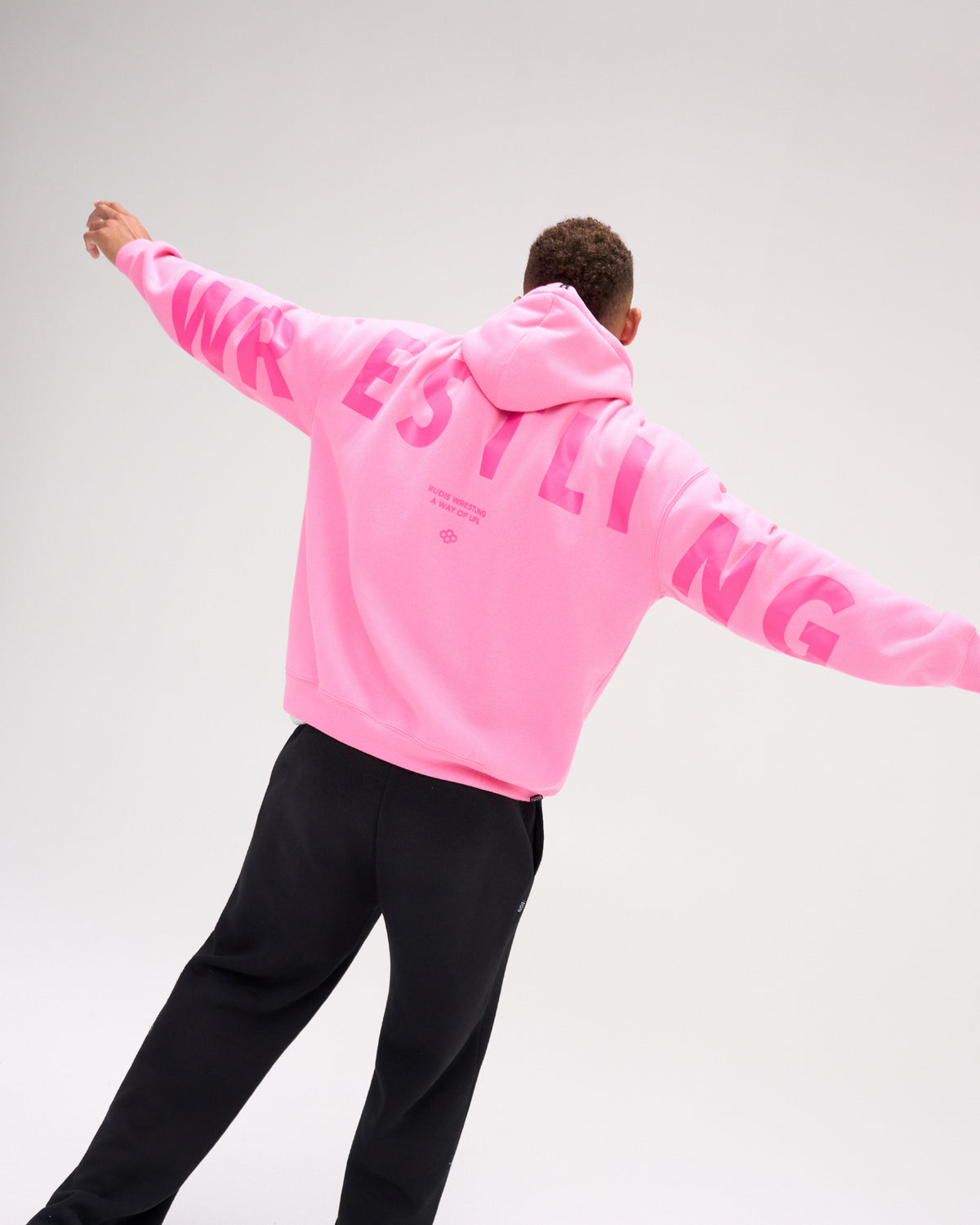 A man wearing a bright pink hoodie with 'WRESTLING' printed in pink across the back, paired with black sweatpants, captured from behind against a light gray studio background.