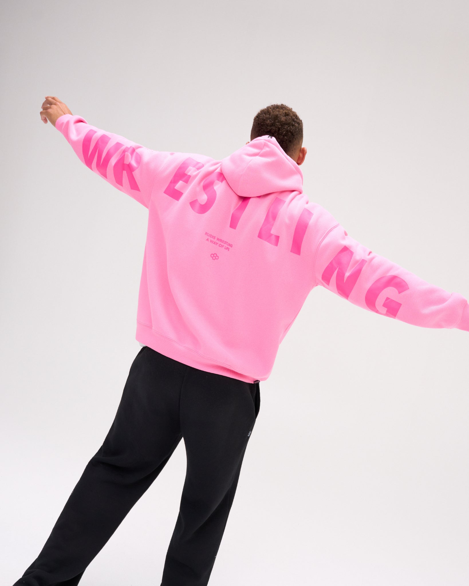 A man wearing a bright pink hoodie with 'WRESTLING' printed in pink across the back, paired with black sweatpants, captured from behind against a light gray studio background.
