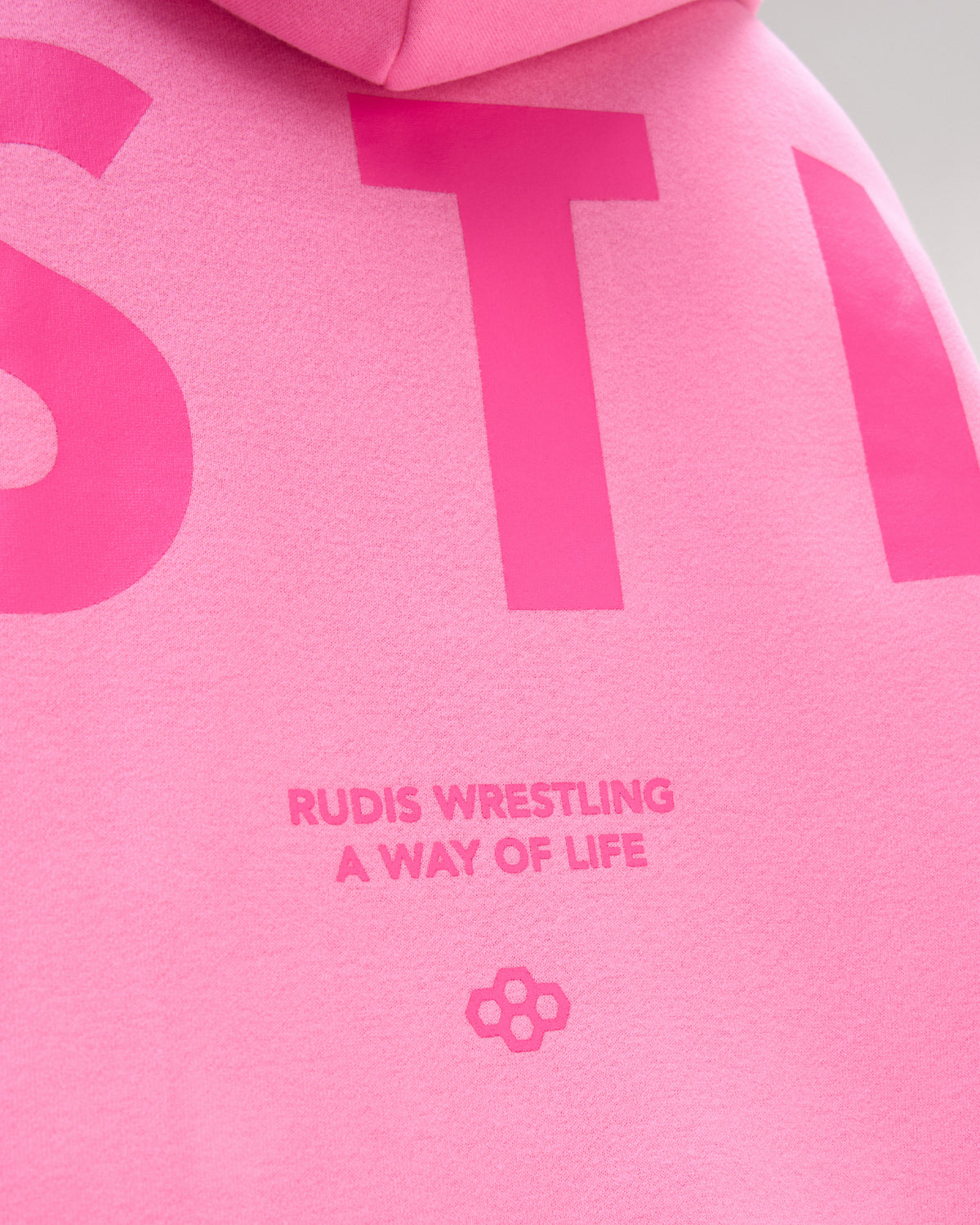 Close-up of a bright pink hoodie showing large tonal pink lettering and the text 'RUDIS WRESTLING A WAY OF LIFE' with a small flower-like logo.