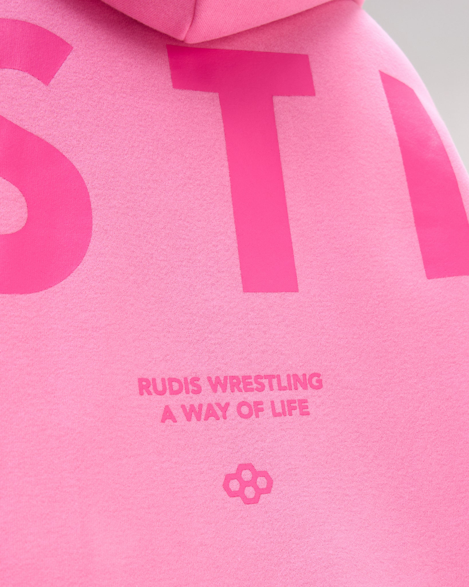 Close-up of a bright pink hoodie showing large tonal pink lettering and the text 'RUDIS WRESTLING A WAY OF LIFE' with a small flower-like logo.