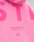 Close-up of a bright pink hoodie showing large tonal pink lettering and the text 'RUDIS WRESTLING A WAY OF LIFE' with a small flower-like logo.