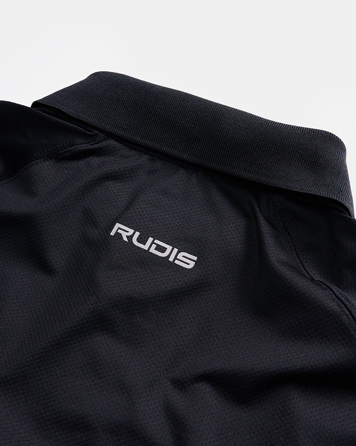 A close-up view of a black athletic polo shirt featuring a slim structured collar and a distinctive logo that reads RUDIS