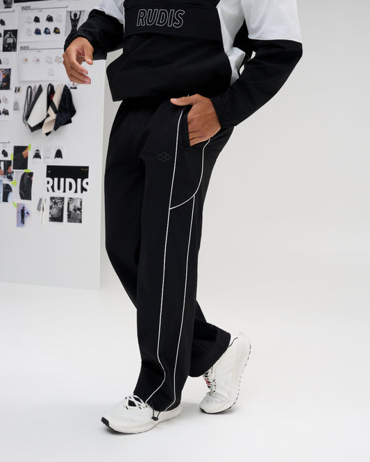 A person modeling black wide-leg track pants with white piping and a small tonal logo, paired with a black-and-white top and white sneakers in a bright studio setting.