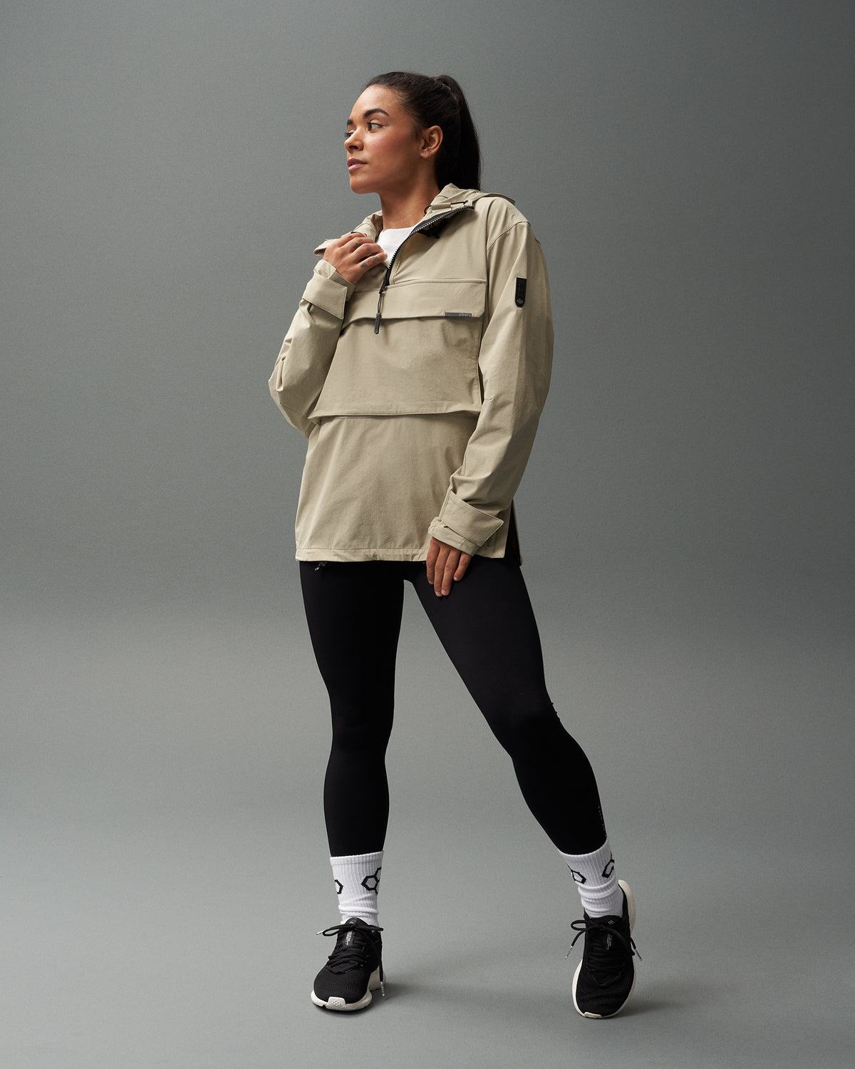 A person wearing a beige 1/4 zip jacket with a hood, paired with black leggings and black sports shoes, standing against a grey background.