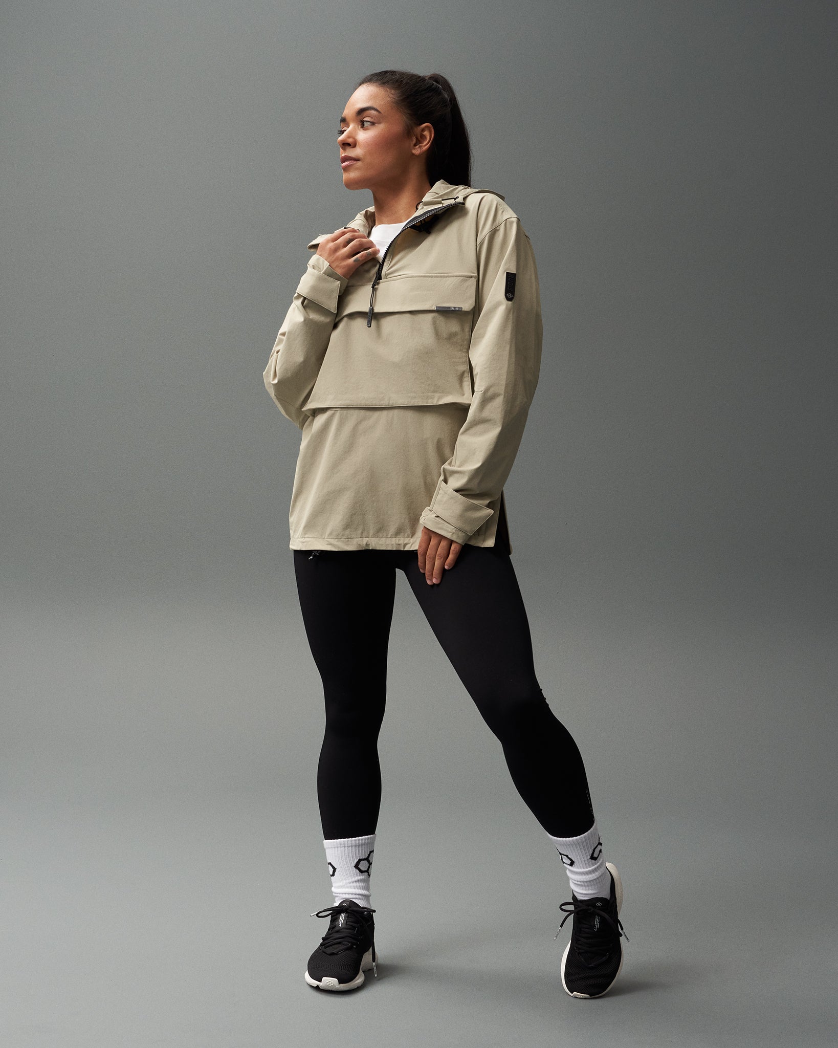 A person wearing a beige 1/4 zip jacket with a hood, paired with black leggings and black sports shoes, standing against a grey background.