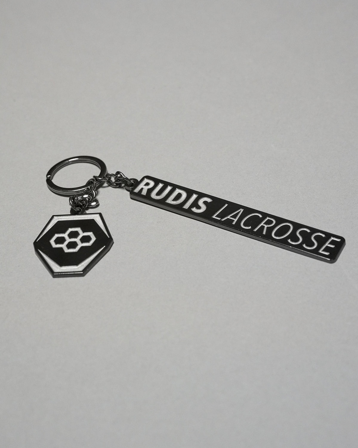RUDIS Lacrosse metal keychain in black and silver featuring the signature RUDIS hex logo, engraved RUDIS Lacrosse branding, durable metal construction, and split key ring.