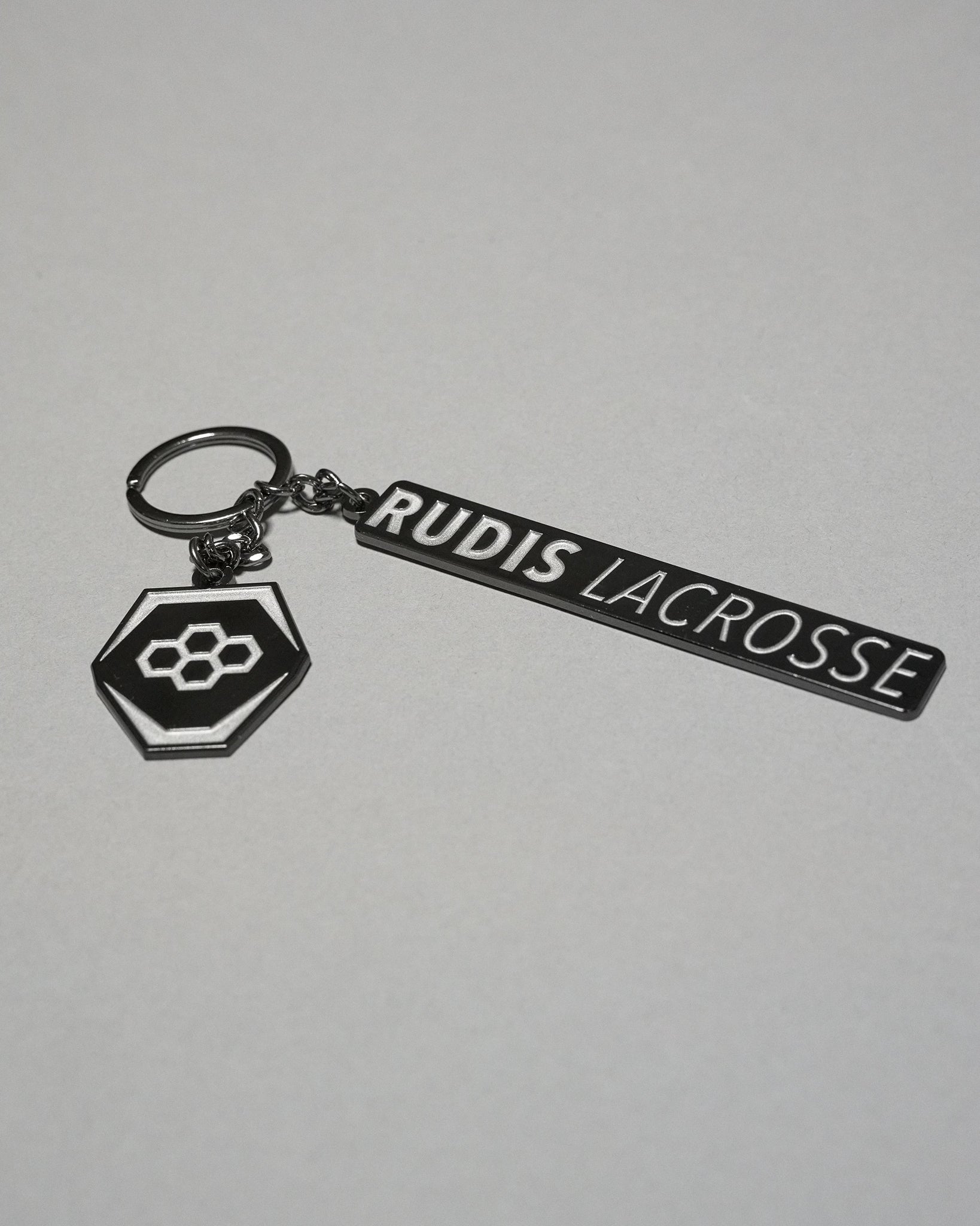 RUDIS Lacrosse metal keychain in black and silver featuring the signature RUDIS hex logo, engraved RUDIS Lacrosse branding, durable metal construction, and split key ring.