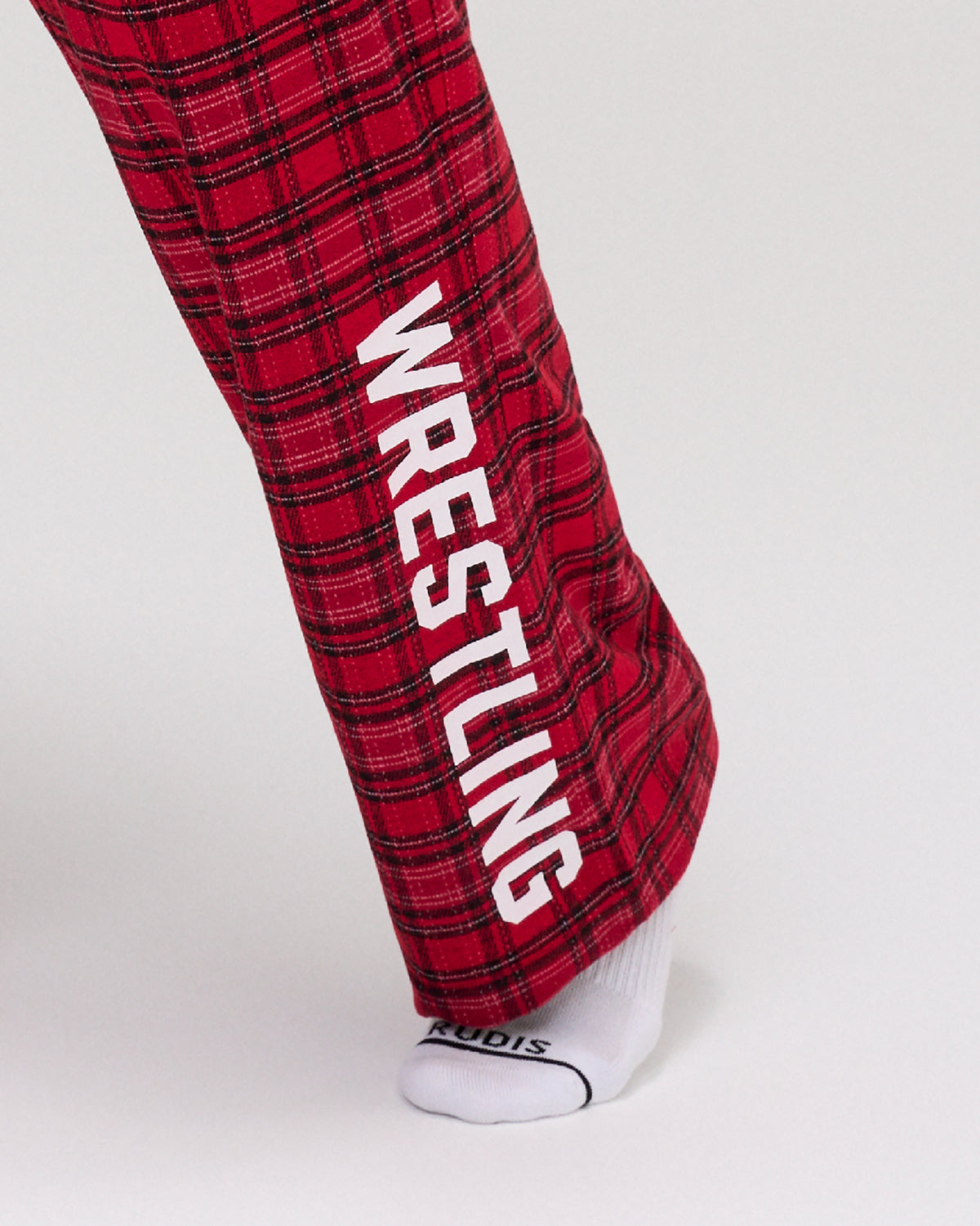 Close-up of RUDIS Hex Flannel Lounge Pants in Red and black plaid, highlighting the large white WRESTLING lettering printed down the lower leg. Copy Alt Text