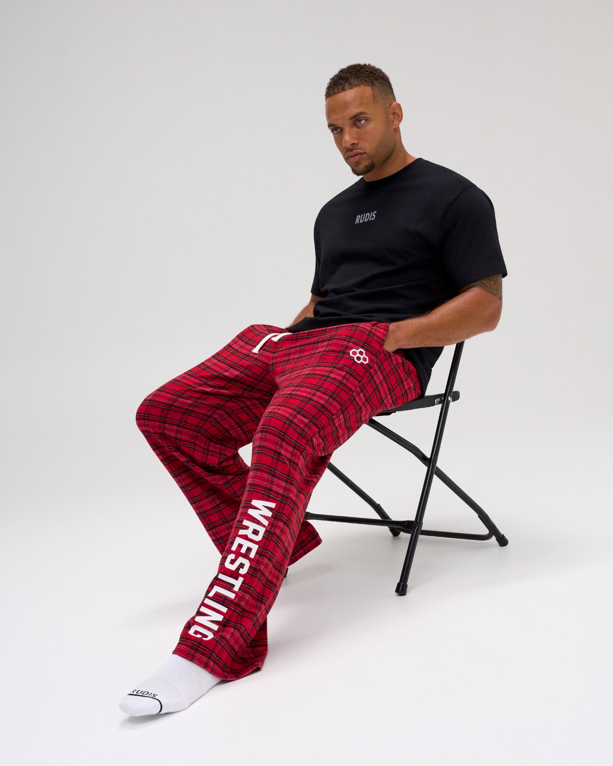 Male model seated in RUDIS Hex Flannel Lounge Pants in Red plaid, showing the relaxed fit, white drawstring waistband and WRESTLING leg graphic. Copy Alt Text