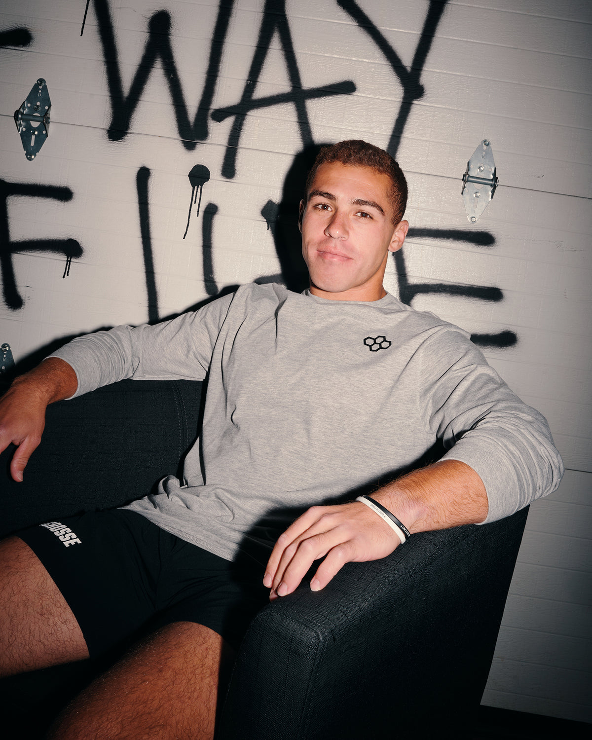 A man wearing a light gray long-sleeve crewneck shirt with a small black logo on the chest, paired with black shorts, sitting in a dark armchair against a white wall with black graffiti.