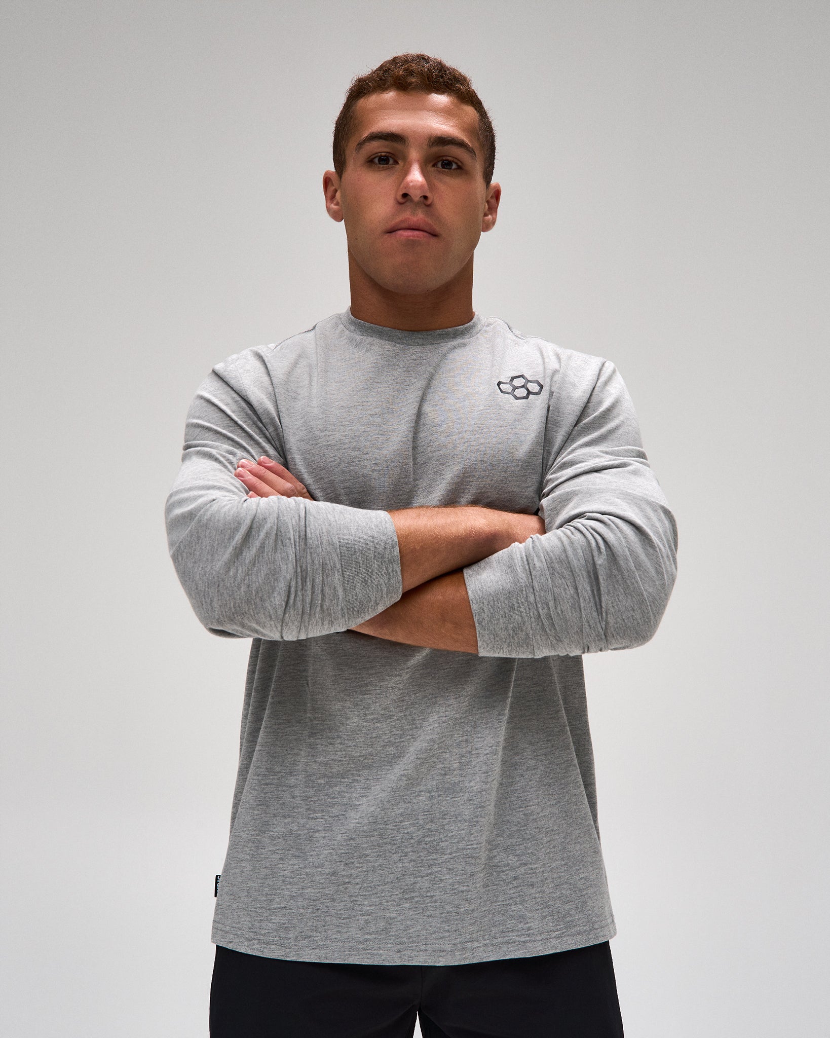 A man wearing a heather gray long-sleeve athletic shirt with a small black logo on the left chest, standing with arms crossed against a light gray studio background.
