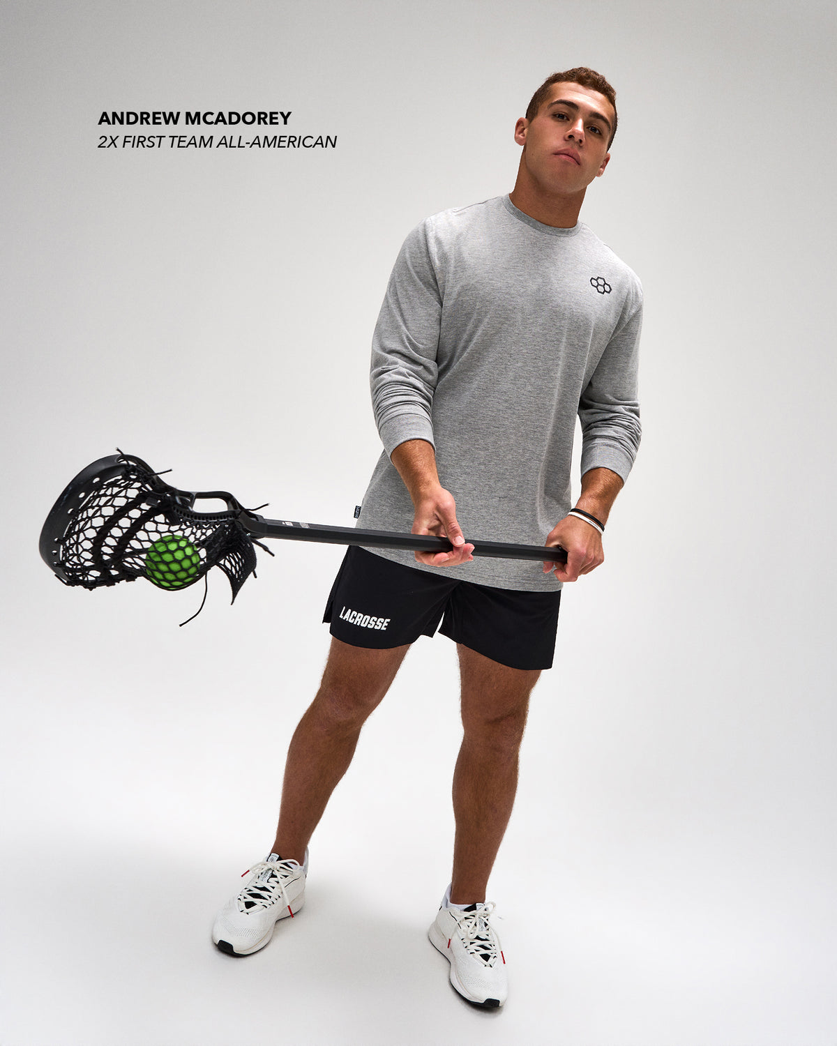 A man modeling athletic apparel, wearing a heather gray long-sleeve Lacrosse shirt, black Lacrosse shorts, and white sneakers, holding a lacrosse stick with a green ball against a light gray studio background.