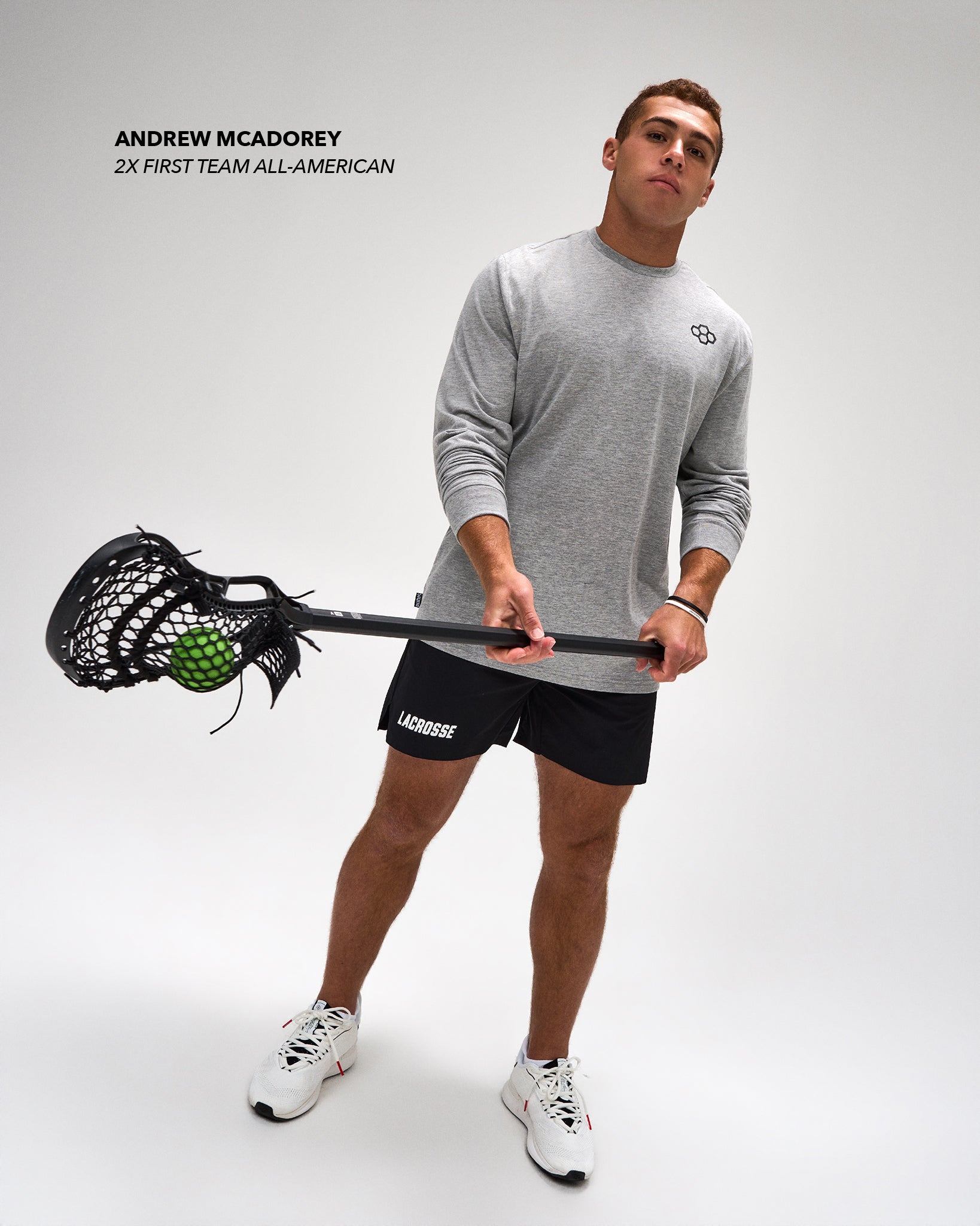 A man modeling athletic apparel, wearing a heather gray long-sleeve Lacrosse shirt, black Lacrosse shorts, and white sneakers, holding a lacrosse stick with a green ball against a light gray studio background.