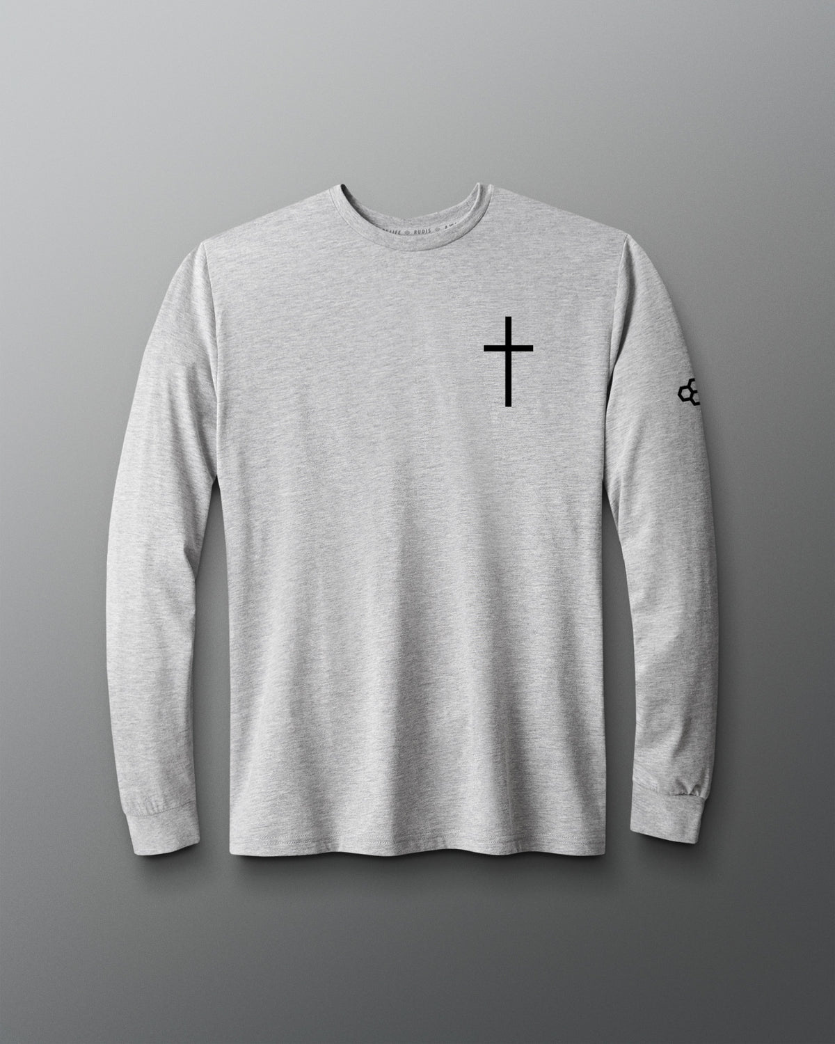 A light gray heather long-sleeve t-shirt with a black cross printed on the left chest and a small black logo on the right sleeve, displayed against a dark gray gradient background.