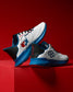 Side view of a pair of white RUDIS training shoes with blue soles, black and red accents, and USA Wrestling and hexagon logos. One heel features the phrase ‘A Way of Life’ in red text. Shoes are displayed on a red background.