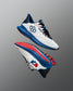 Pair of white RUDIS training shoes with blue midsoles, red and black detailing, and hexagon and USA Wrestling logos. The outsoles feature a red tread pattern. Shoes are displayed floating against a red-to-blue gradient background.