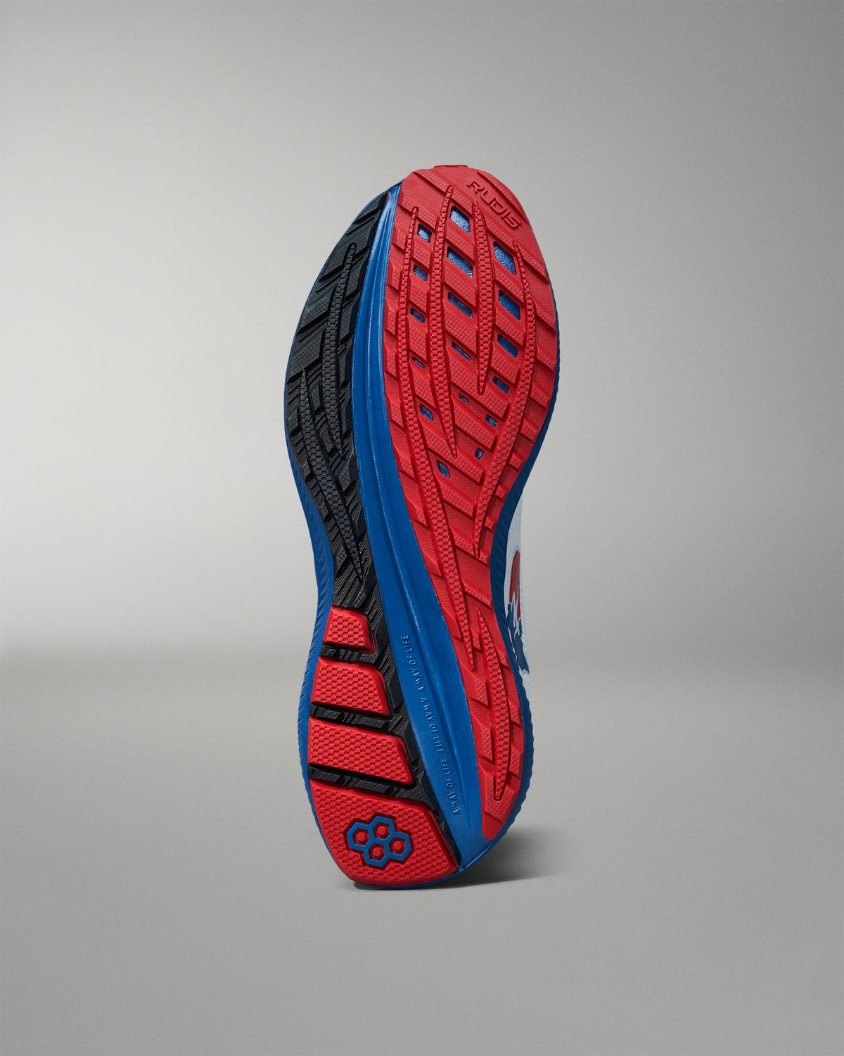 Bottom view of a RUDIS training shoe showcasing a red, blue, and black high-traction outsole with a hexagon logo near the heel. Displayed against a neutral gray background.