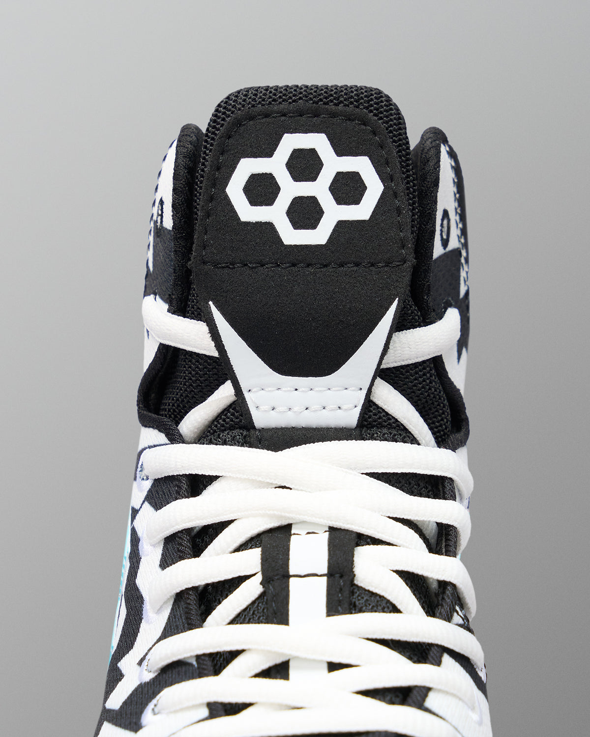 Close-up of a high-top wrestling shoe showing the black tongue with a white geometric logo, white laces, and white contrast overlays on a gray background.