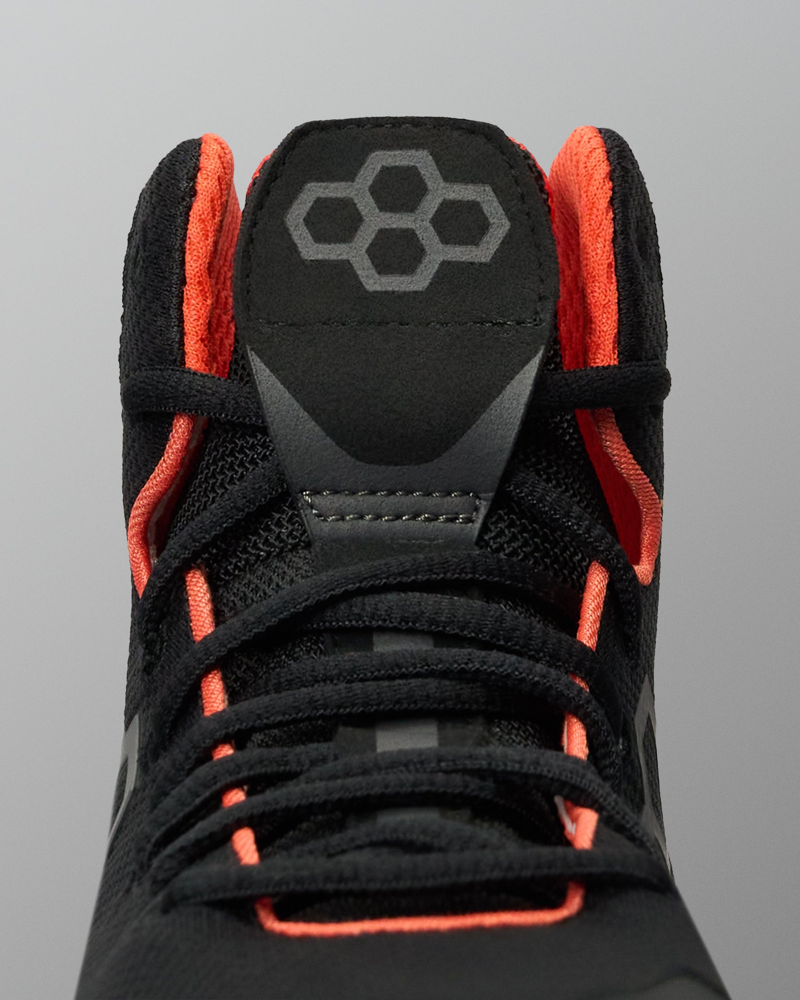Close-up of a black Wrestling shoe with bright orange lining and laces, featuring a gray geometric logo on the tongue, set against a solid gray background.