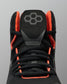Close-up of a black Wrestling shoe with bright orange lining and laces, featuring a gray geometric logo on the tongue, set against a solid gray background.