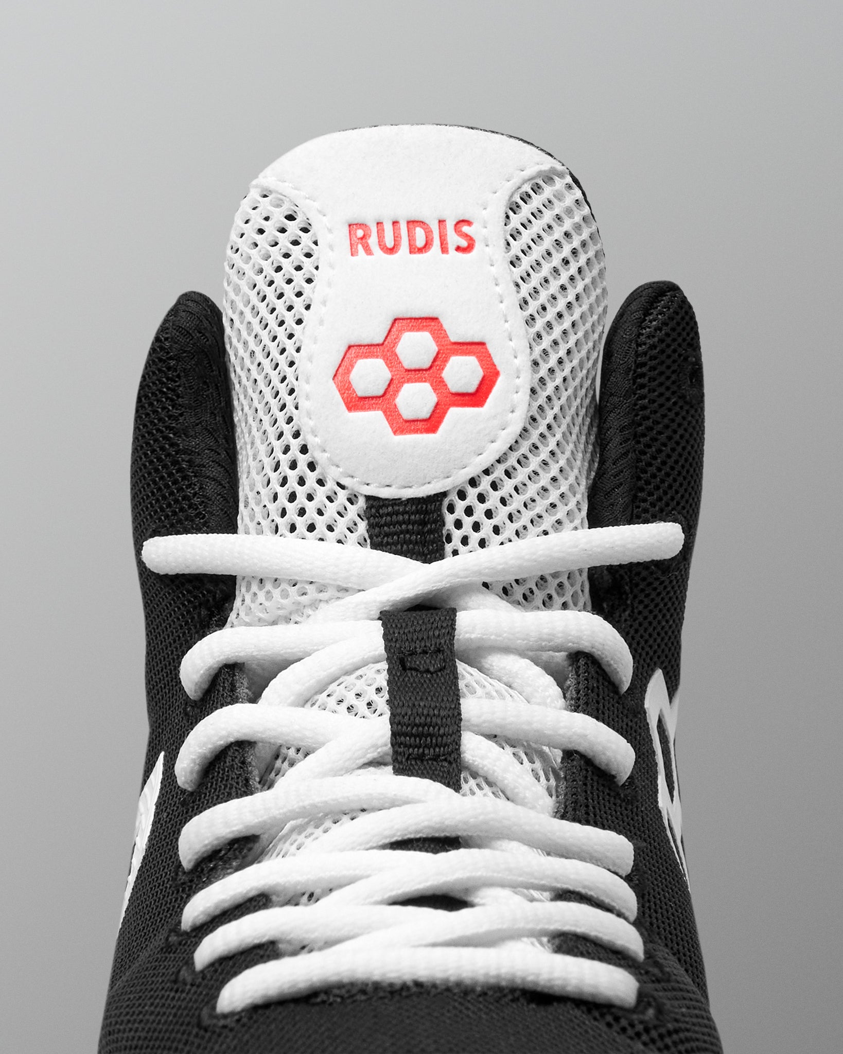 Close-up of a black and white high-top wrestling shoe showing the tongue with the brand name RUDIS in red and a red geometric logo, white laces, and a gray background.