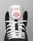 Close-up of a black and white high-top wrestling shoe showing the tongue with the brand name RUDIS in red and a red geometric logo, white laces, and a gray background.