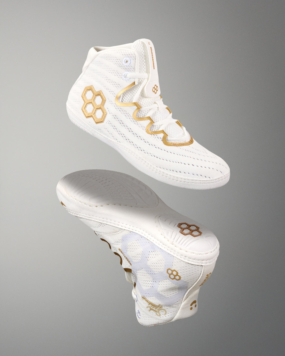 A pair of white wrestling shoes with gold quadhex and wavy geometric logo details, shown floating against a neutral gray gradient background.
