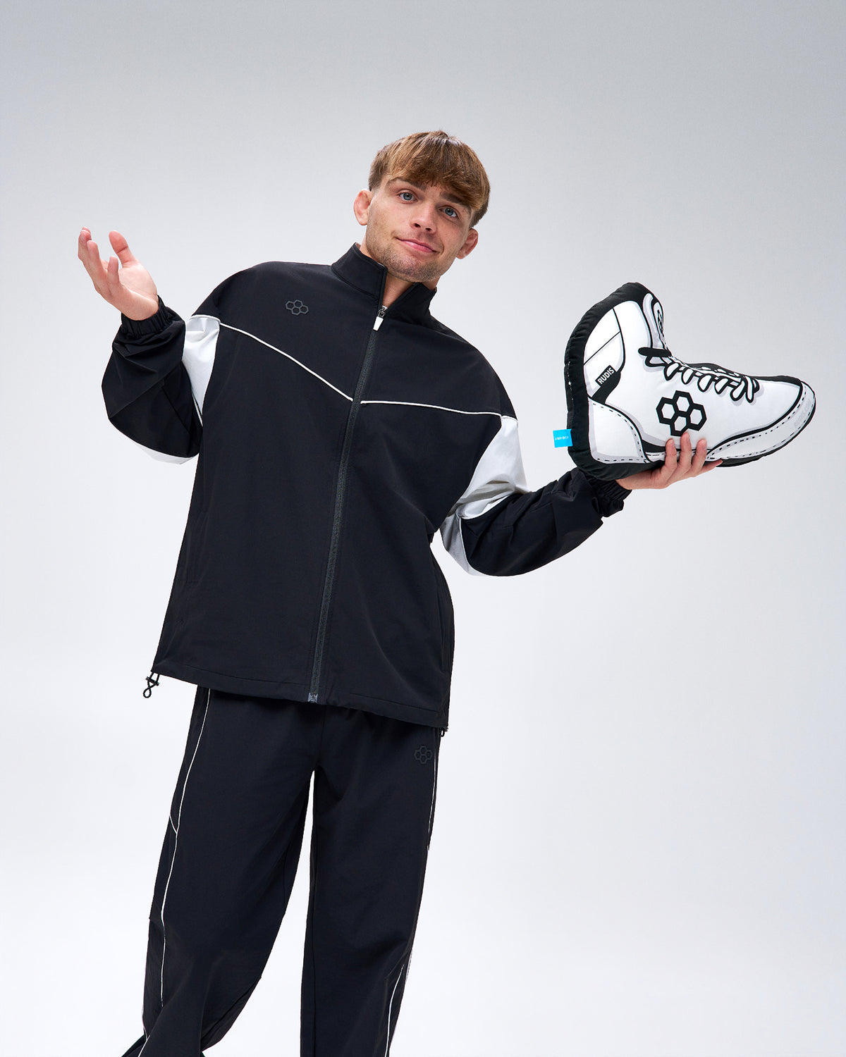 Model wearing a black RUDIS tracksuit holding the RUDIS Colt Shoe 18-inch Pillow in White, featuring a wrestling shoe design with black laces and RUDIS branding.