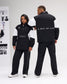 A man and a woman modeling matching black and white color-block tracksuits in a bright studio setting. The woman is shown from the front wearing a black half-zip jacket with white shoulder panels and 'RUDIS' printed on the chest, paired with black pants.