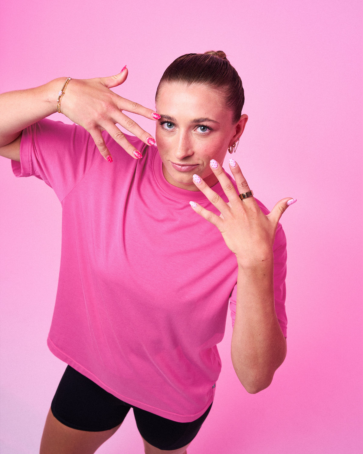 A woman posing in a bright pink athletic t-shirt and black shorts against a solid pink studio background.