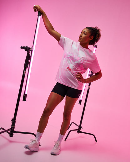 A woman posing in a pink studio setting wearing a light pink graphic t-shirt, black athletic shorts, white socks, and white sneakers, holding a vertical light bar on a stand.