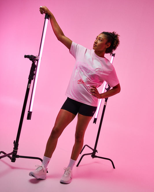 A woman posing in a pink studio setting wearing a light pink graphic t-shirt, black athletic shorts, white socks, and white sneakers, holding a vertical light bar on a stand.