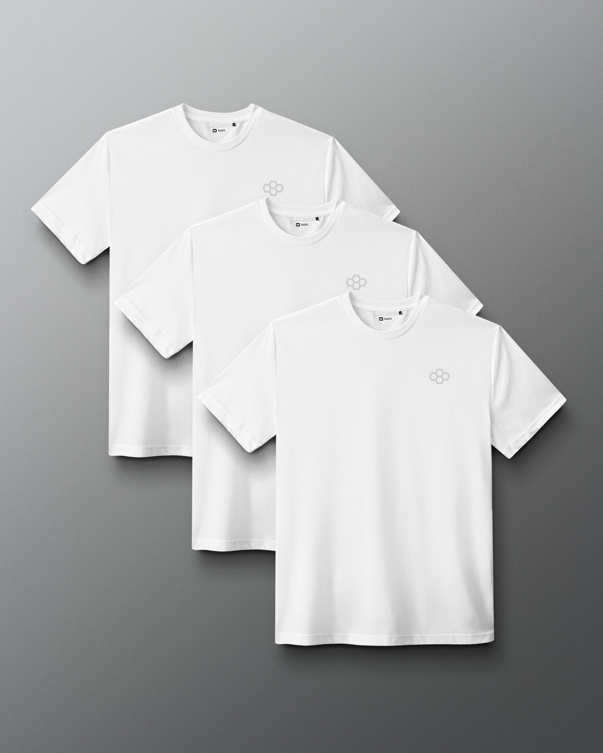 Three white short-sleeve crewneck t-shirts with a small gray geometric logo on the left chest, arranged in a staggered overlapping layout on a dark gray gradient background.
