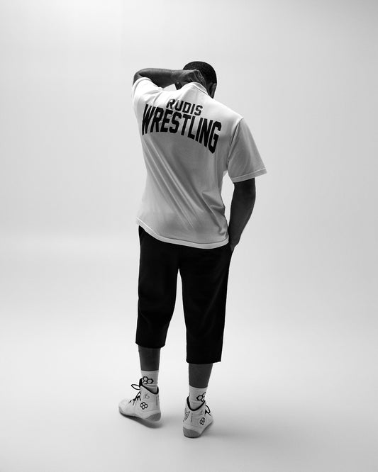 Back view of the white RUDIS All In Boxy Hybrid T-Shirt featuring bold black RUDIS WRESTLING lettering, styled with black pants and wrestling shoes.