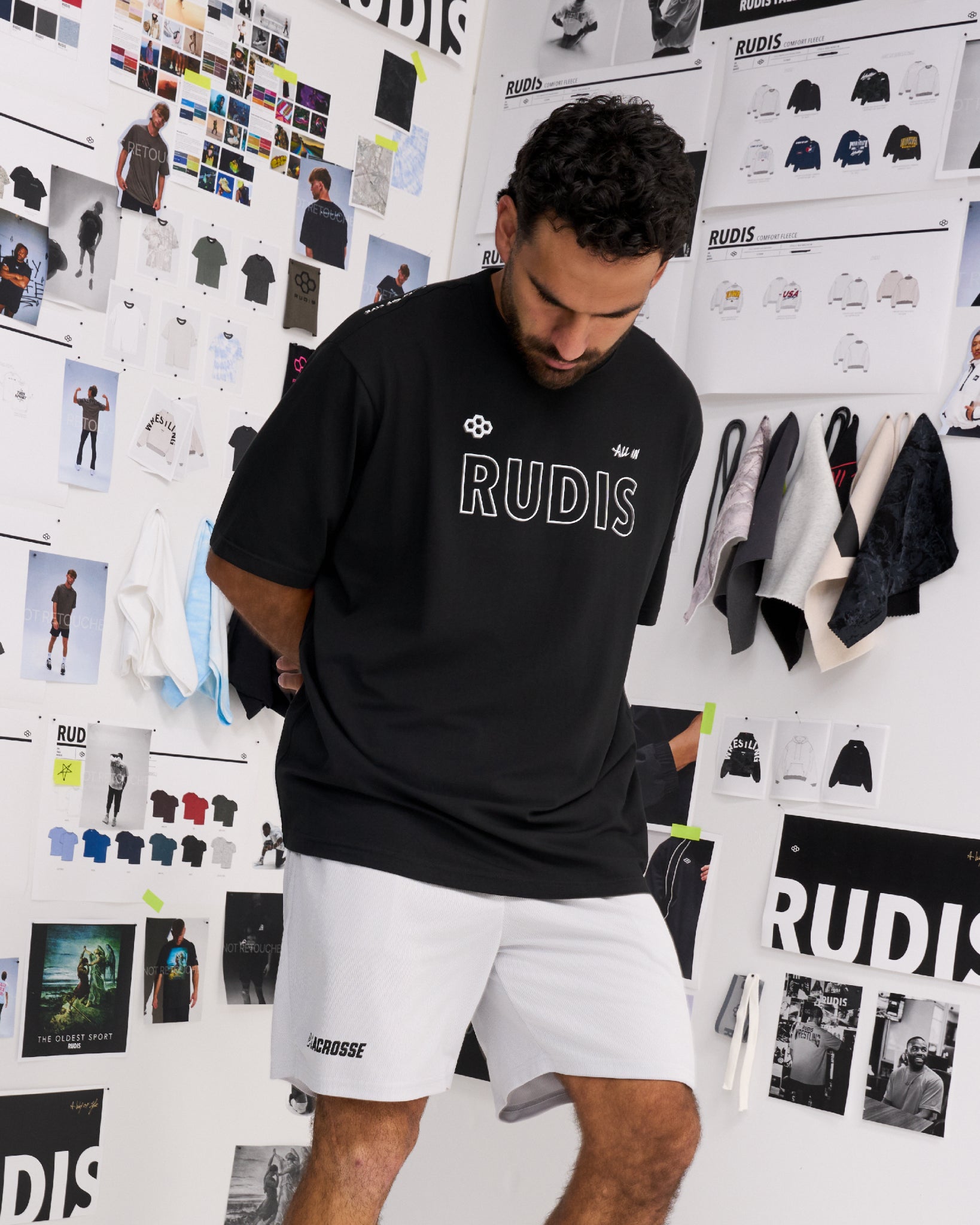 A man wearing a black RUDIS t-shirt and white shorts, standing in front of a white wall covered with fashion design sketches and apparel displays.