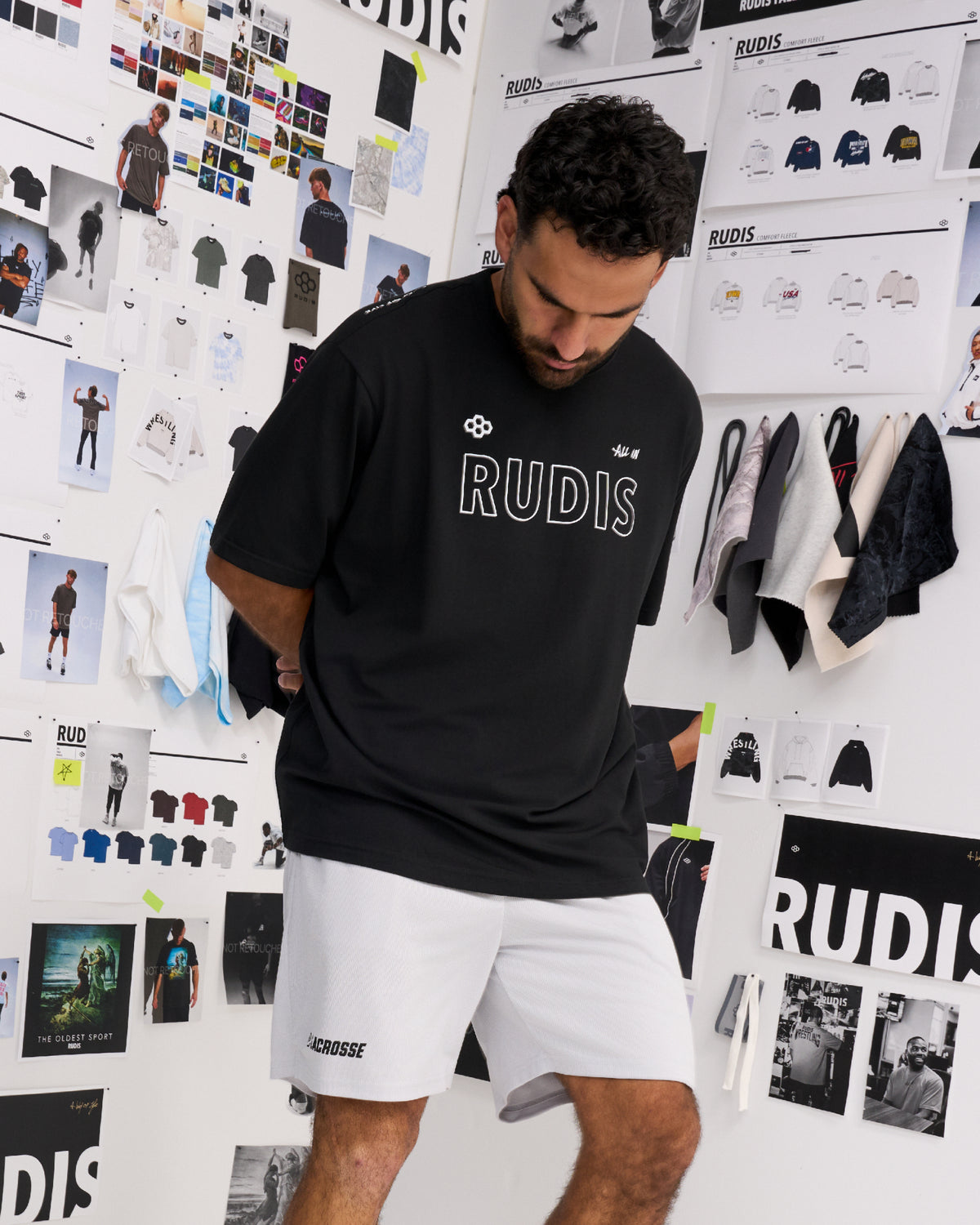 A man wearing a black RUDIS t-shirt and white shorts, standing in front of a white wall covered with fashion design sketches and apparel displays.